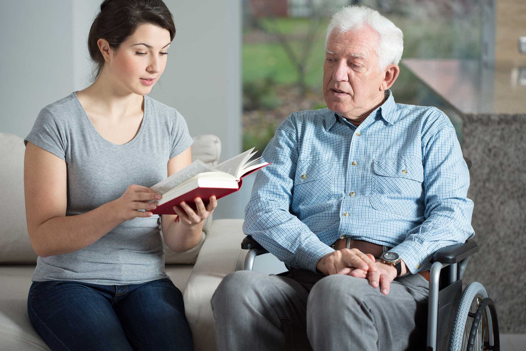 Female caregiver reading a book to an elderly man in a wheel chair